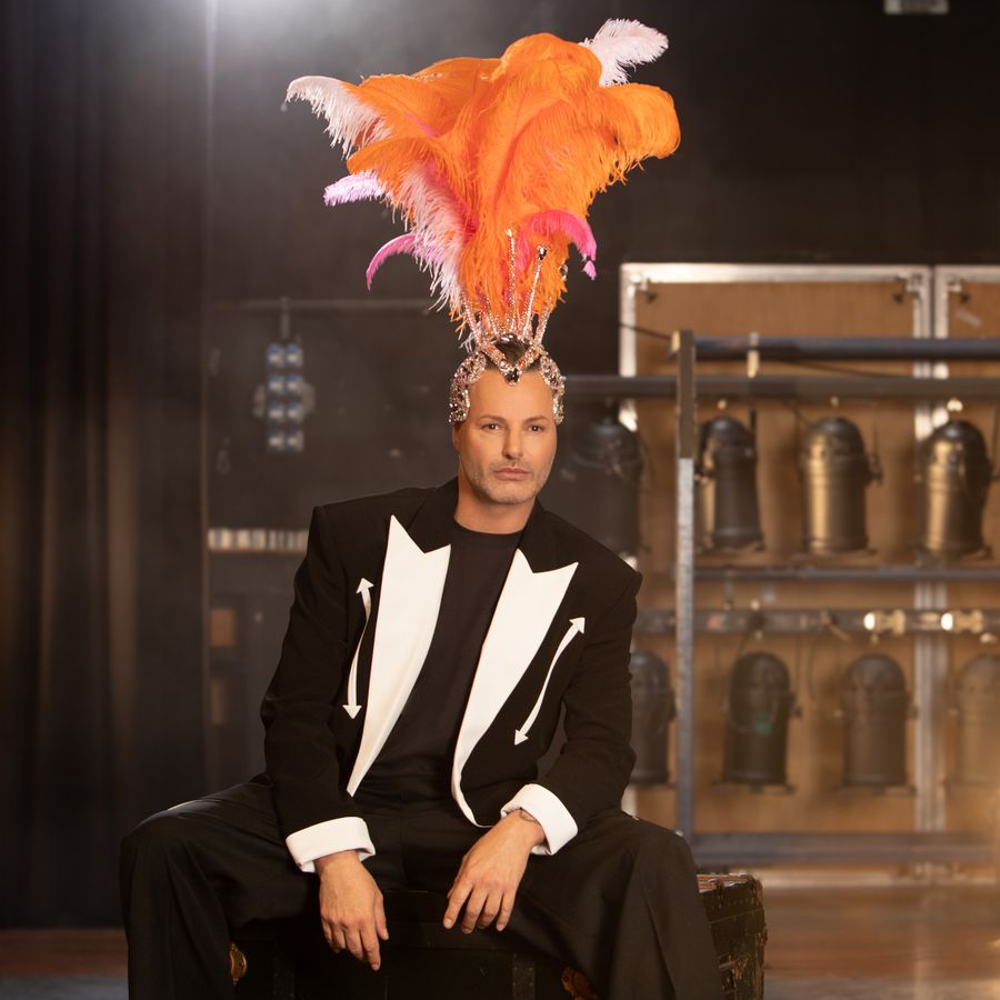 Man in chique smokingpak met witte accenten zit op toneel met op achtergrond theaterlampen in een rek. Op zijn hoofd draagt hij een verentooi welke showgirls dragen. 