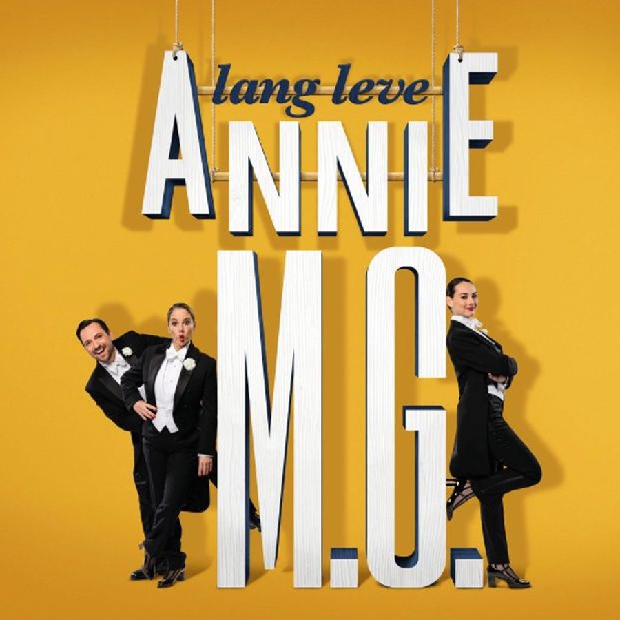 Een geel plaatje met witte letters: lang leve Annie M.G. met drie mensen in een zwart pak. Twee vrouwen en een man.