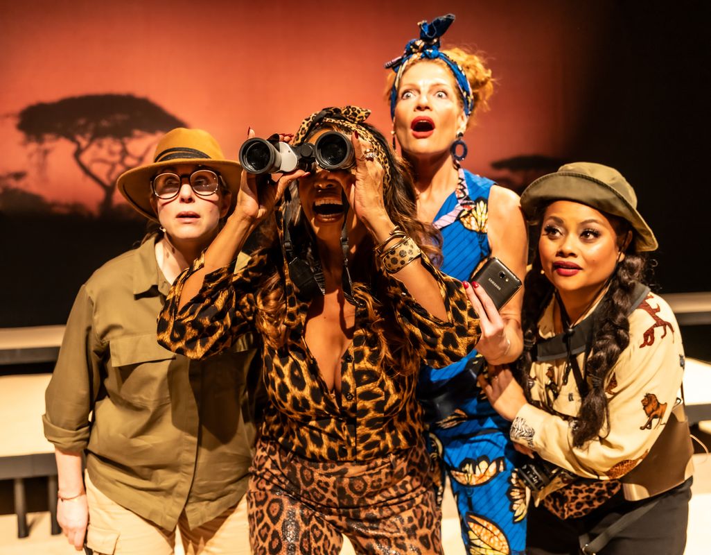 4 dames in safari outfits waarvan er eentje door een verrekijker kijkt en de rest ook in dezelfde richting kijkt. 