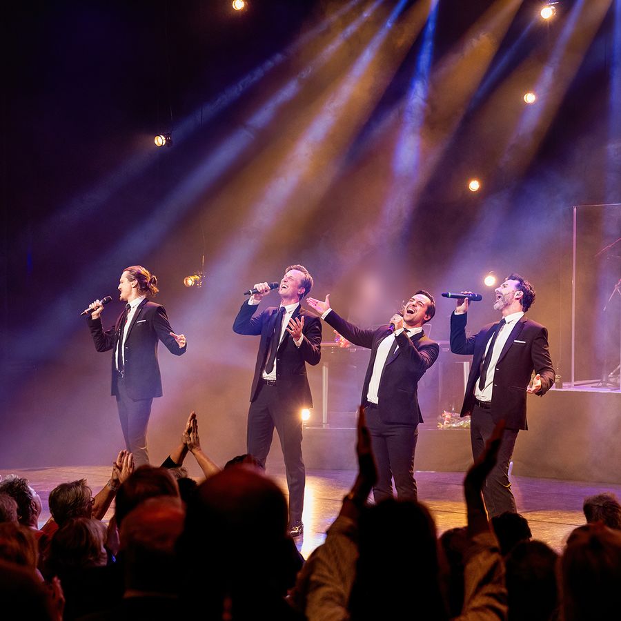 4 mannen in zwarte smokings staand op de voorrand van een mooi uitgelicht podium, overtuigend zingend.