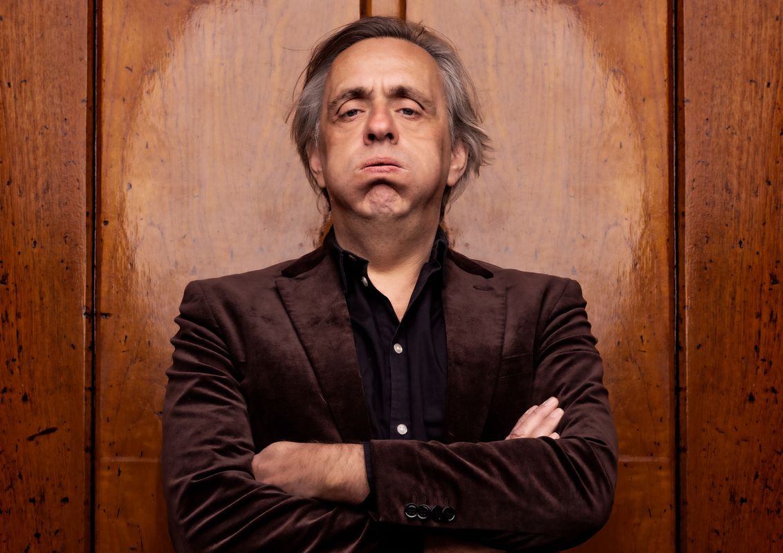 Marcel van Roosmalen heeft zijn wangen opgeblazen en kijkt naar de camera. Hij kijkt niet heel blij. Op de achtergrond is hout te zien met een lichtkring achter zijn hoofd. Hij heeft zijn armen over elkaar heen geslagen