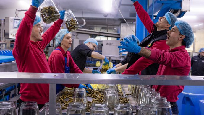 Vier personen in rode overall met blauwe haarnetjes over hun haar staan aan de lopende band van een augurkenfabriek. Een houdt twee potten met augurken de lucht in. 