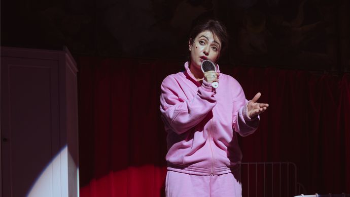 Een vrouw in een roze trainingspak met een korte broek staat met een haarborstel in haar handen alsof ze een microfoon vast heeft. Achter haar staat een bed en een kast en op de achtergrond is er een rood doek te zien.
