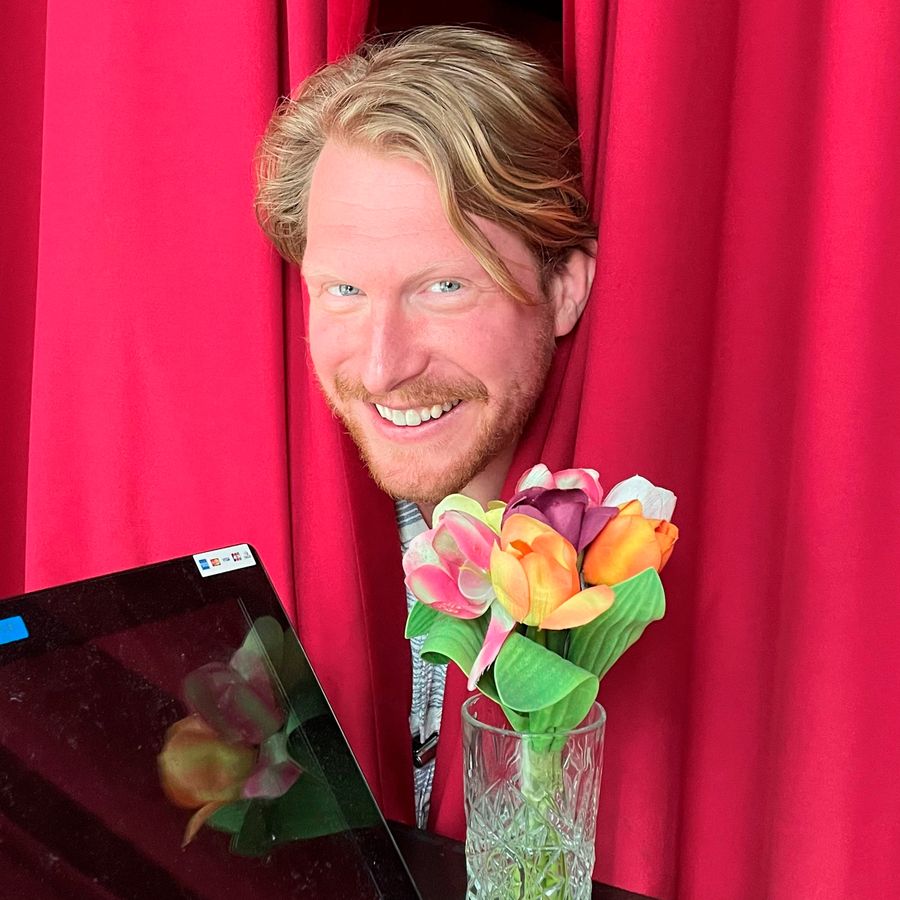 Het hoofd van Kor Hoebe verschijnt tussen een roze theaterdoek, met bloemen onder zich.