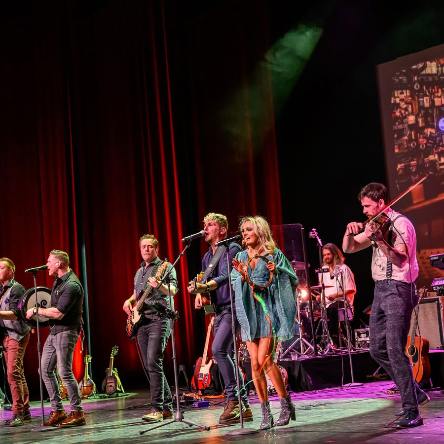 Een 9 koppige band met diverse instrumenten met als middelpunt een dame in een blauwe groene jurk met tamboerijn. 