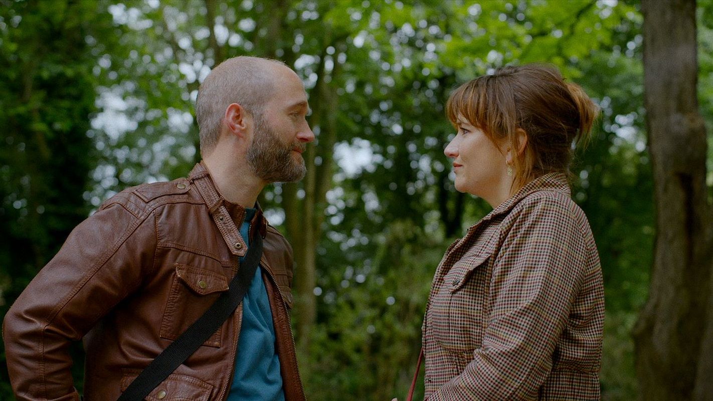 Man met bruin leren jasje staat tegen over vrouw in een bos. 