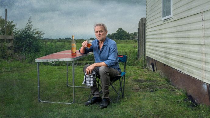 Marcel zit naast een muurtje in een soort weiland aan een campingtafeltje op een campingstoel. Hij houdt een theedoek in zijn handen en drinkt uit een glas. Op tafel naast hem staat een fles drank. Hij kijkt ernstig de camera in.