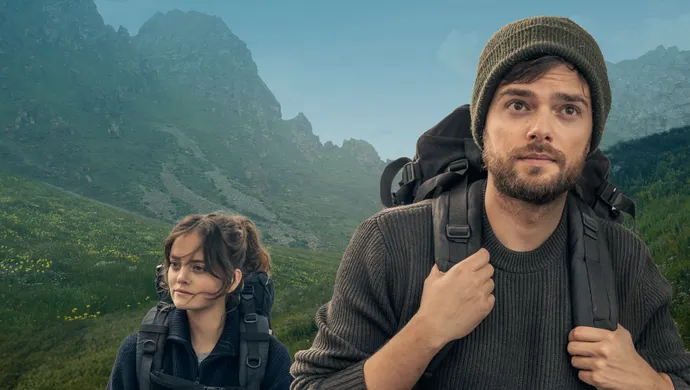 Man met wandelrugzak op de voorgrond kijkend in de camera,  achter hem een een vrouw gekleed in blauw, ook met wandelrugzak, wandelend door een groen bergachtig gebied. 