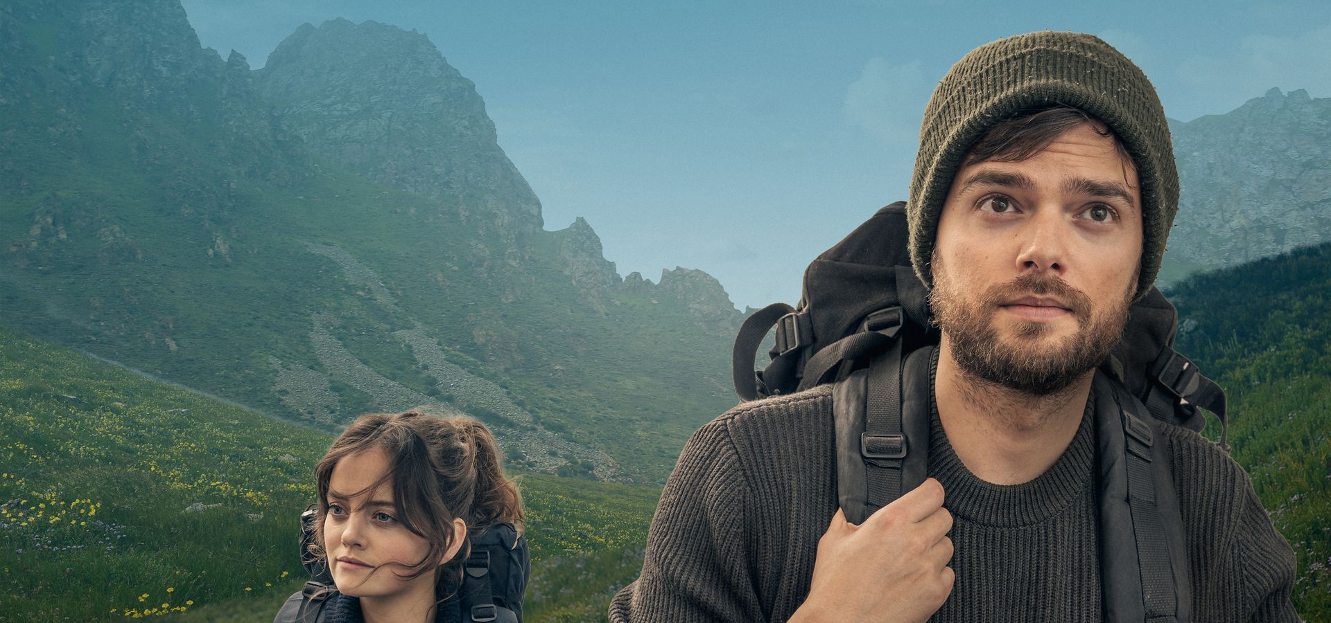 Man met wandelrugzak op de voorgrond kijkend in de camera,  achter hem een een vrouw gekleed in blauw, ook met wandelrugzak, wandelend door een groen bergachtig gebied. 