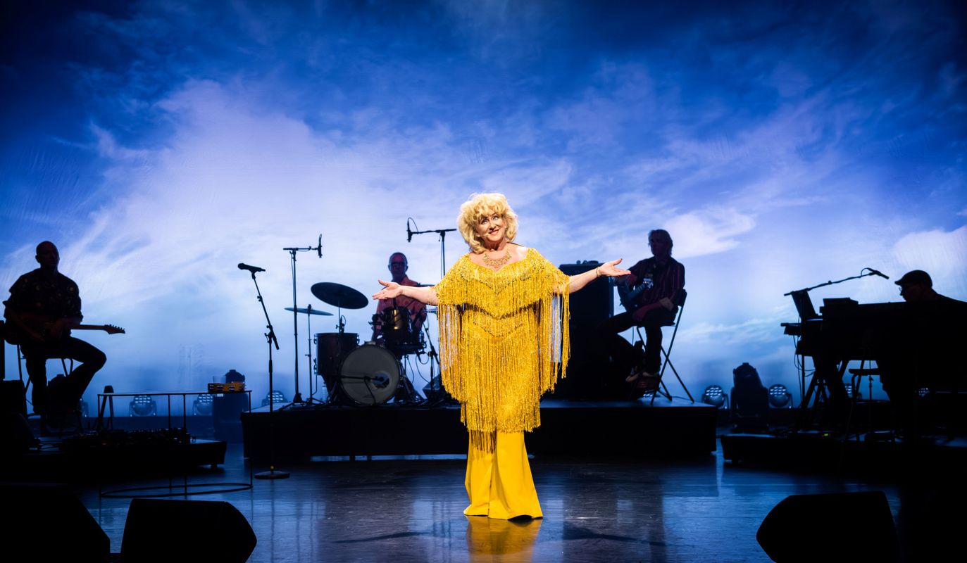 Karin Bloemen staat in een gele jurk met haar handen wijd op een podium. Achter haar staat een band en is ere een blauwe kleur te zien.
