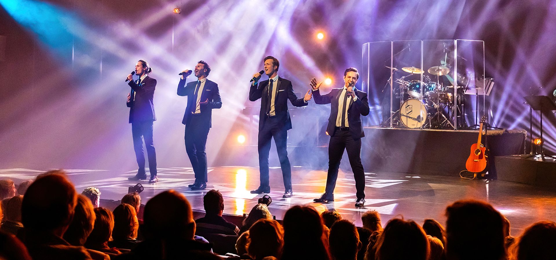 4 mannen in zwarte smokings staand op de voorrand van een mooi uitgelicht podium, overtuigend zingend.
