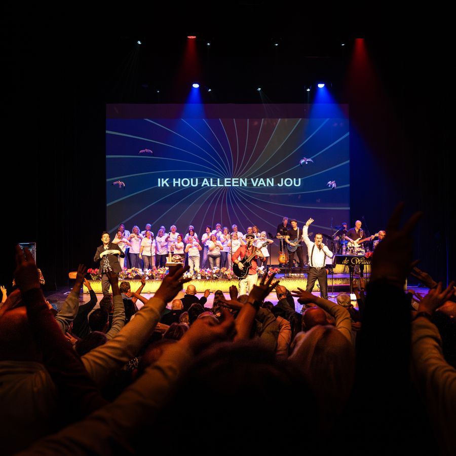Een podium met allemaal mensen erop. De foto is gemaakt vanuit het publiek en handen steken in de lucht. 