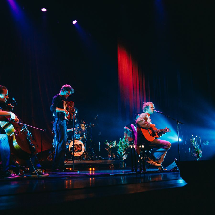 Er staat een band op een podium. Een man met een gitaar zit op een krukje voor een microfoon en zingt. De achtergrond is donker en er hangt een rood theatergordijn.
