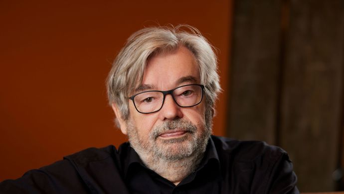 Maarten van Rossem kijkt serieus naar de camera en zit op een bank. Hij heeft zijn handen over elkaar.