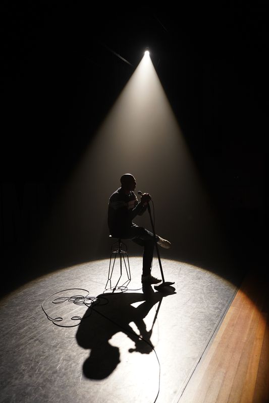 Man zittend op een stoel op een geheel donker podium met één spotlight op hem gericht.  De schaduw van zijn profiel zichtbaar op de grond. 