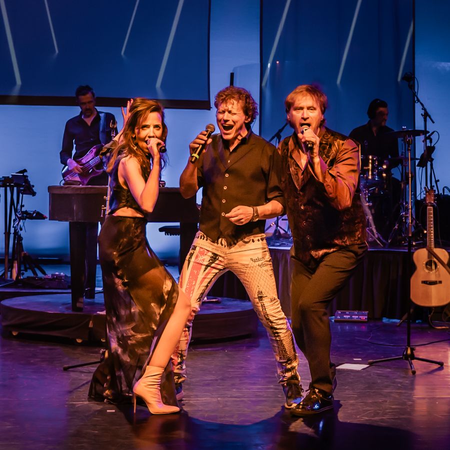 Twee mannen en een vrouw staan center stage en houden een microfoon vast en zingen erdoorheen. Op de achtergrond zie je een deel van de band