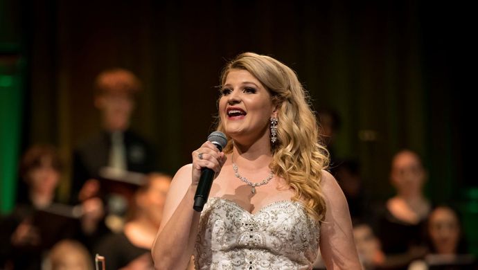 Een vrouw in een witte jurk houdt een microfoon vast en zingt. Op de achtergrond staat een koor.
