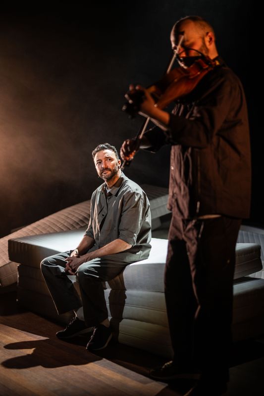 Man zitten op een stapel matrassen kijkt op naar een man die viool speelt. 