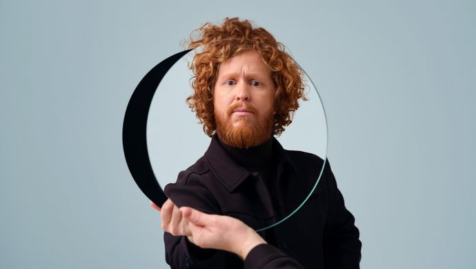 Man in zwart en bos krullen houdt zichzelf een spiegel voor. 