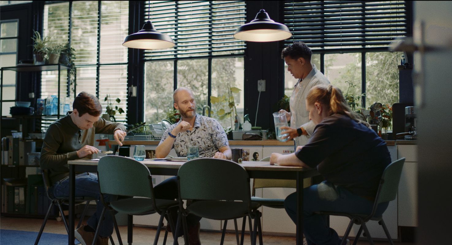 Vier personen zittend aan een lange tafel. Twee daarvan kijken naar hun bord, eentje kijkt op naar iemand die aan komt lopen met een kan water. 
