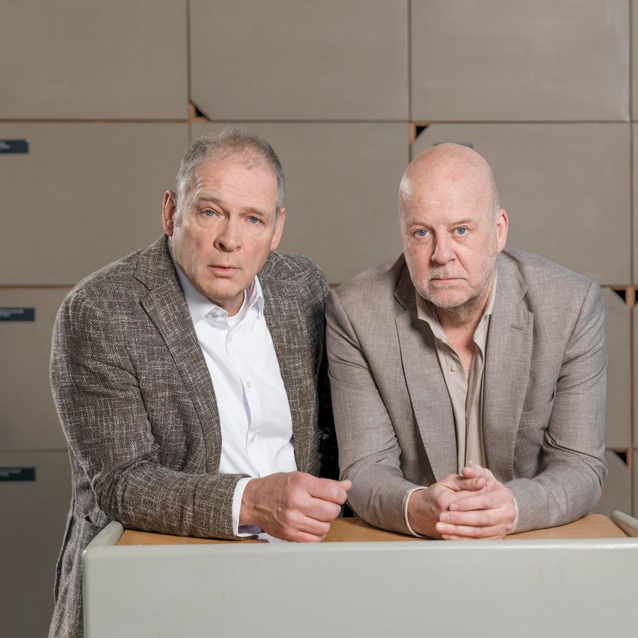 Twee mannen, kalend in grijze/beige jasjes leunen op een tafel, achter hen een rij lockerkastje. 