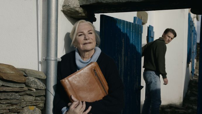 Een vrouw met tas in de armen staat in het zonlicht voor haar huis, met op de achtergrond een jonge man die de deur dicht doet en naar haar kijkt. 