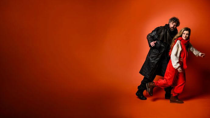 Een rood geklede vrouw en een zwart geklede man rennen en kijken verschrikt achterom. De achtergrond is oranje.