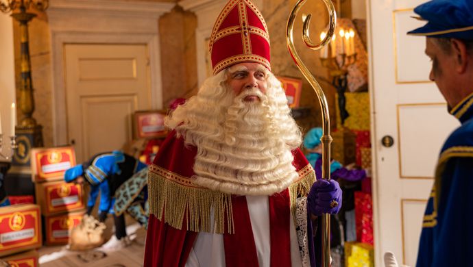 Sinterklaas met staf in de hand kijkt naar iemand in de deuropening. 