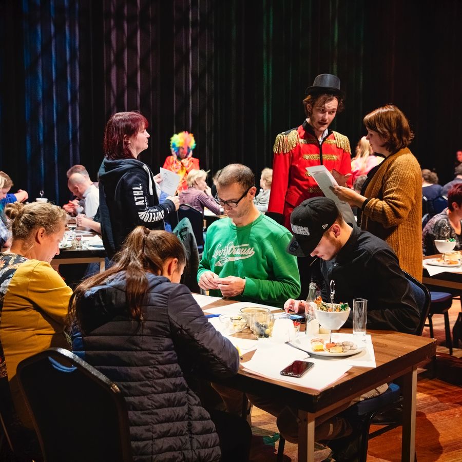 Er zitten allemaal groepjes mensen aan tafel met allemaal papieren op tafel. Midden in de groep loopt een man rond in een rood pak met een zwarte hoge hoed.