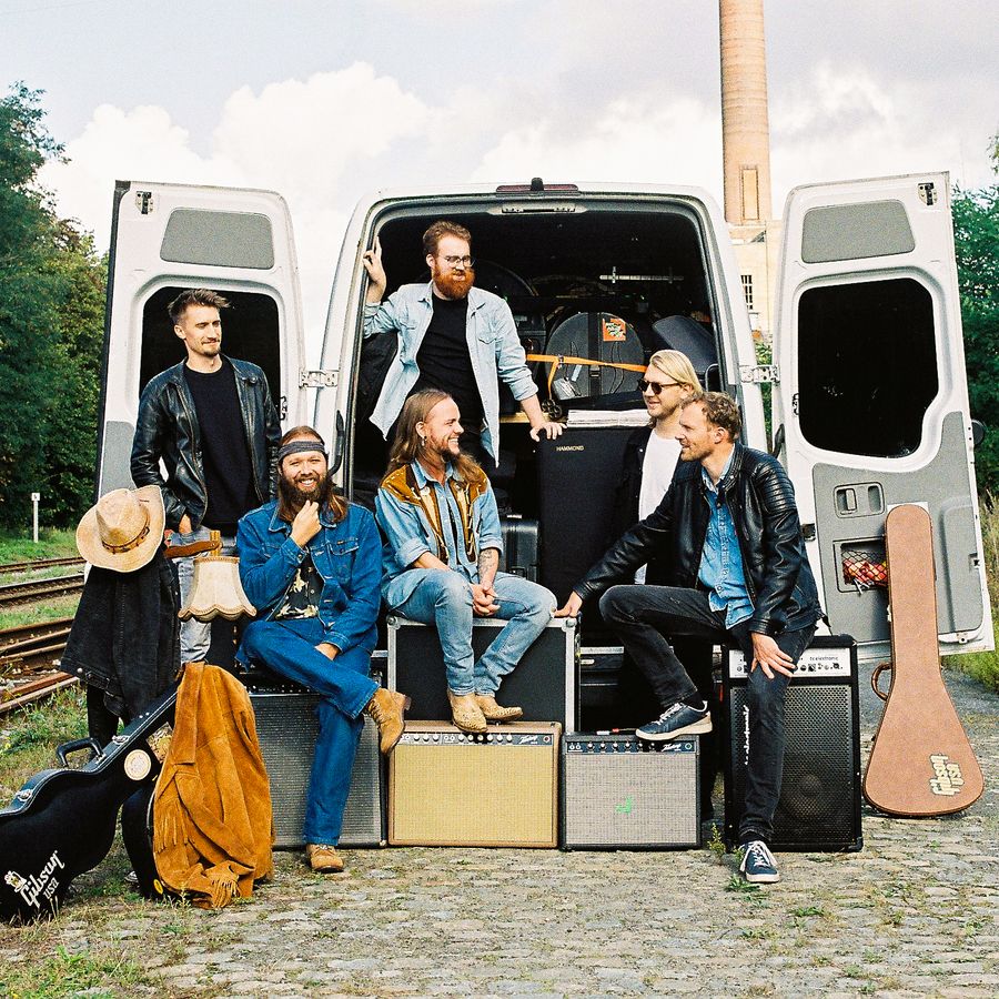 6 koppige band zit met instrumenten en spullen voor een wit busje waarvan de deuren open zijn. 
