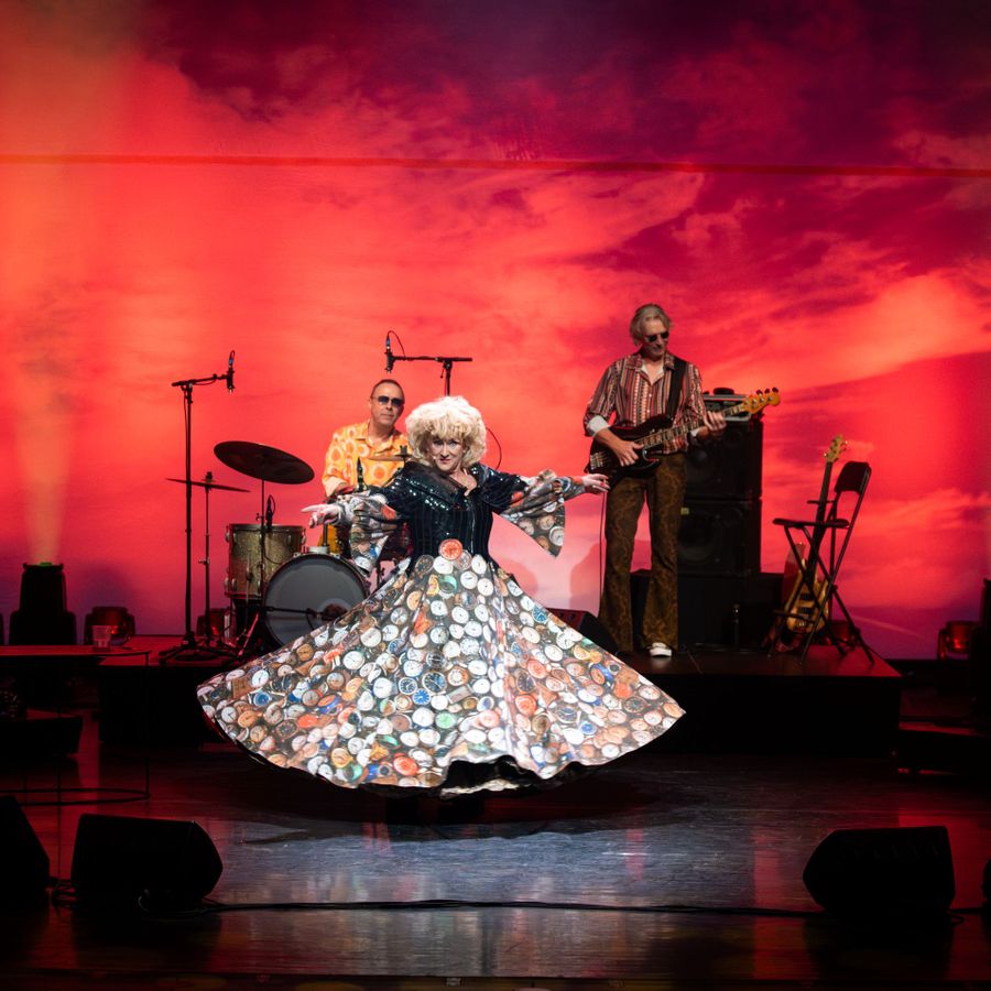 Karin Bloemen staat met een grote jurk op het podium met een band achter haar. Ze heeft haar handen wijd en kijkt uitdagend het publiek in. Op de achtergrond is een rode kleur te zien.