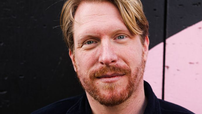 Kor Hoebe staat voor een zwarte achtergrond met een beetje roze. Hij heeft een zwart shirt aan en houdt zijn handen op zijn rug. Hij kijkt glimlachend de camera in.