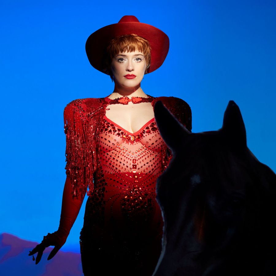 Er staat een vrouw en een paard voor een blauwe achtergrond. De vrouw heeft een rode top aan met gaas zodat je haar buik ziet en ze heeft een rode hoed op.