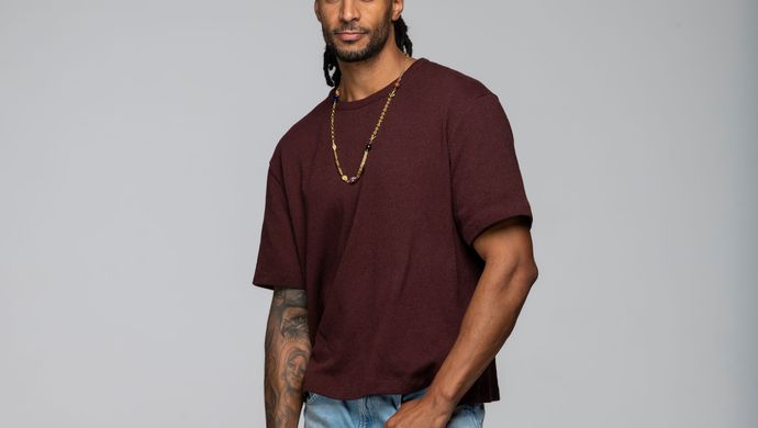 Jay staat voor een witte achtergrond in een spijkerbroek en een donkerrood shirt. hij heeft een hand los langs zijn lijf hangen en de andere hand bij zijn broekzak. Hij kijkt neutraal de camera in.