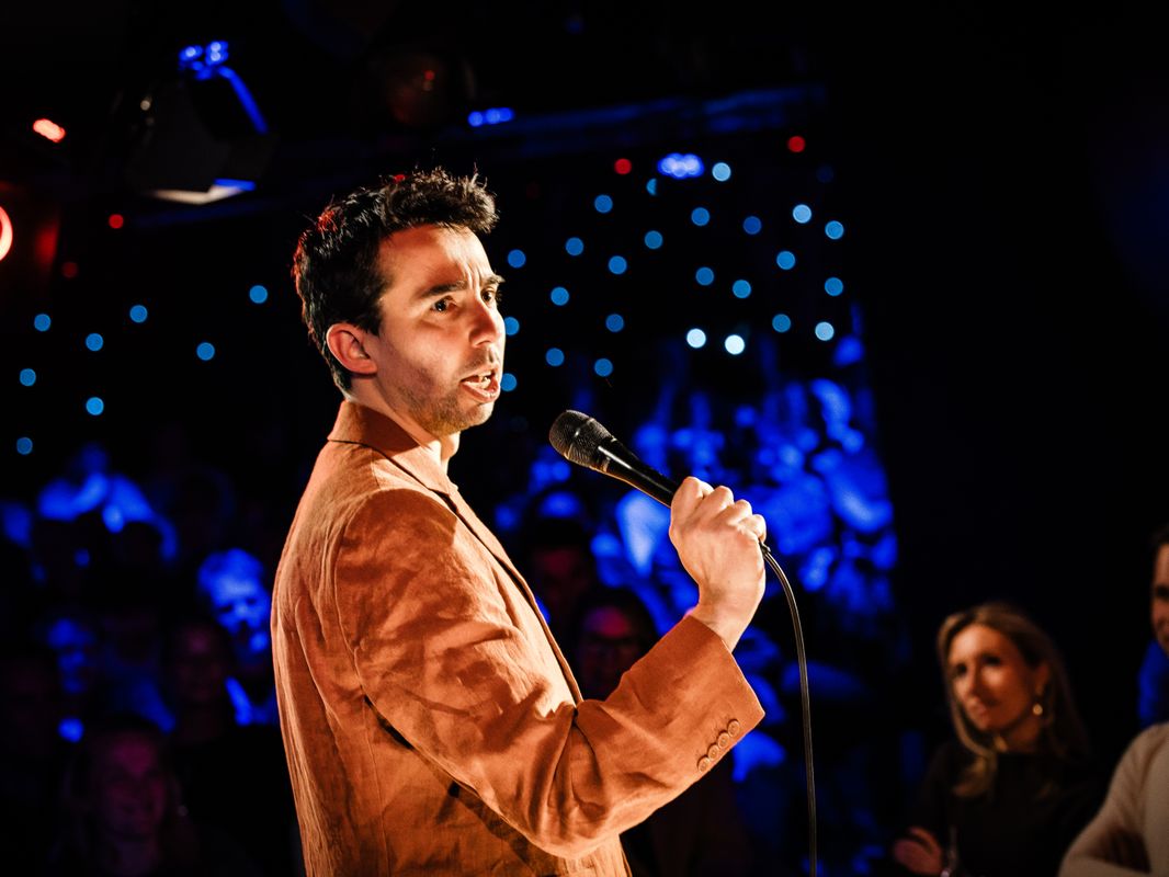 Man met suede bruin jasje houdt een microfoon vast en kijkt naar het publiek. 