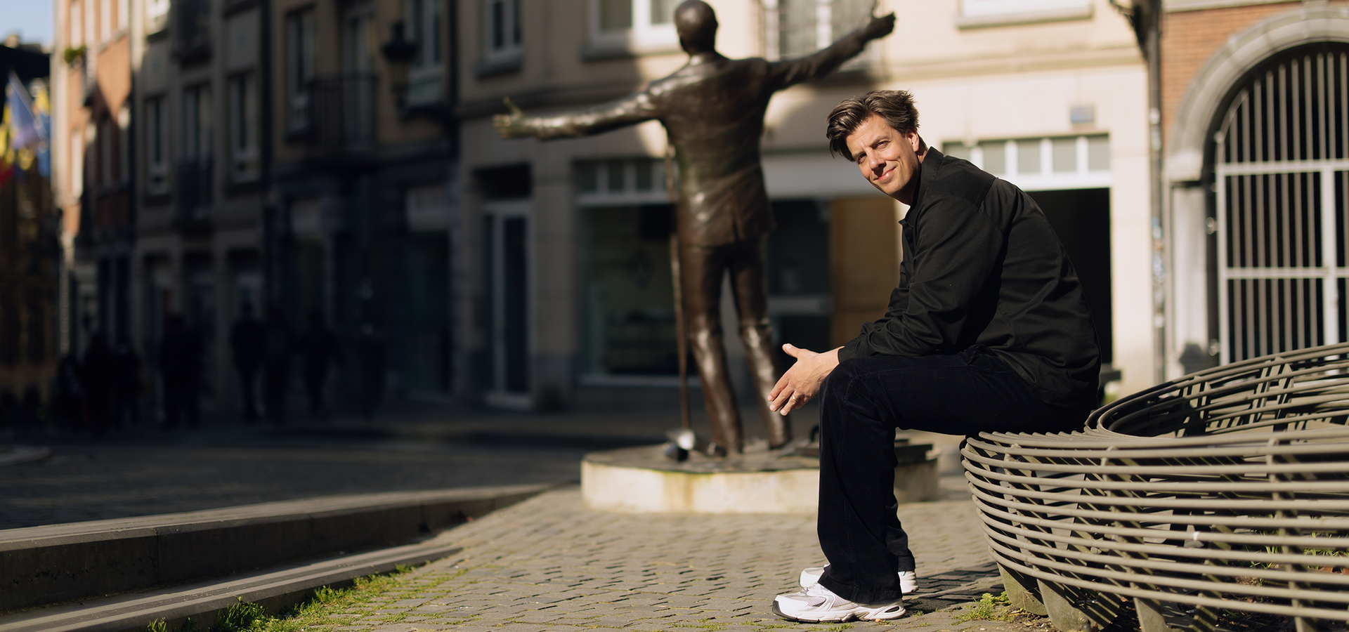 Rob kemps zit voorovergebogen op een bankje met zijn handen bij elkaar. De omgeving is een pleintje met een standbeeld in het midden. Hij kijkt glimlachend de camera in terwijl hij opzij kijkt.