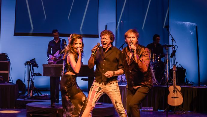 Twee mannen en een vrouw staan center stage en houden een microfoon vast en zingen erdoorheen. Op de achtergrond zie je een deel van de band