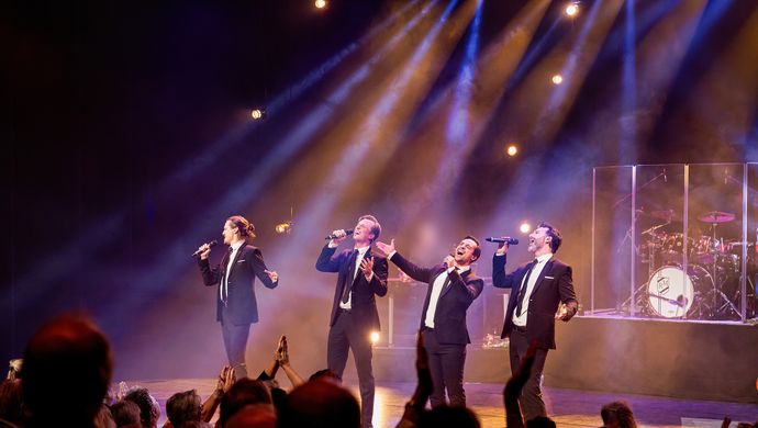 4 mannen in zwarte smokings staand op de voorrand van een mooi uitgelicht podium, overtuigend zingend.