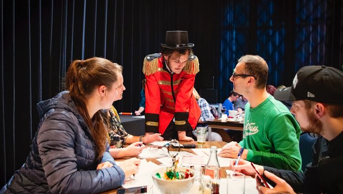 Vier mensen zitten aan een tafel met allemaal papieren. Een man in een rood pak met een zwarte hoed op buigt over de tafel. De mensen aan de tafel kijken allemaal naar de man.