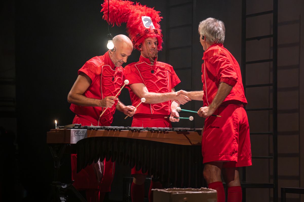 Drie mannen in rode pakjes staan bij een instrument waar ze met stokjes op slaan. Een man schudt de hand van een andere man en staan tegenover elkaar. De derde man staat ernaast en kijkt naar het instrument.