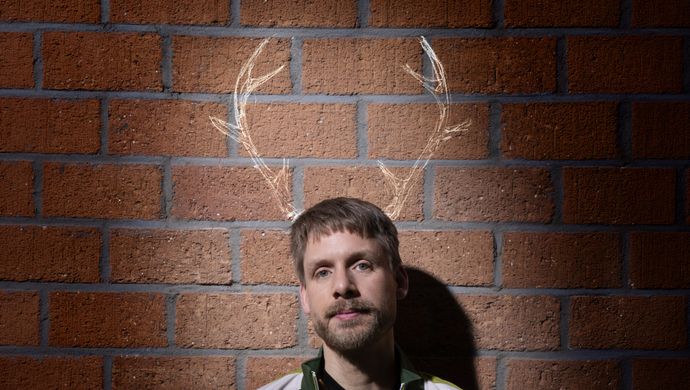 Thijs staat voor een bakstenen muur en kijkt neutraal de camera in. Boven zijn hoofd zijn rendiertakken getekend op de muur.