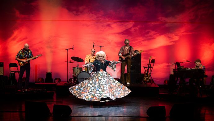 Karin Bloemen staat met een grote jurk op het podium met een band achter haar. Ze heeft haar handen wijd en kijkt uitdagend het publiek in. Op de achtergrond is een rode kleur te zien.