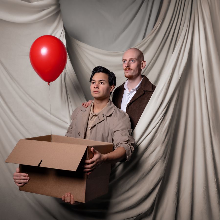 Twee mannen, waarvan een kaal en de ander met donker bruin haar staan achter elkaar. De voorste houdt een verhuisdoos vast en een rode ballon. 