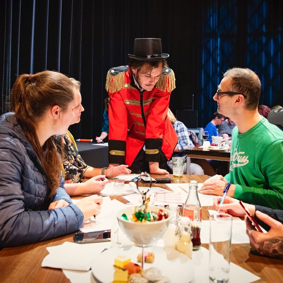 Vier mensen zitten aan een tafel met allemaal papieren op tafel. Ze kijken naar een man in een rode pak en een zwarte hoed op die voorovergebogen naast de tafel staat mee te kijken.