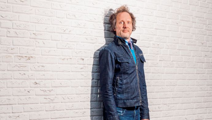 Ronald staat tegen een witte muur. Hij kijkt serieus naar de camera en staat met zijn armen langs zijn lijf, stijf tegen de muur aan.