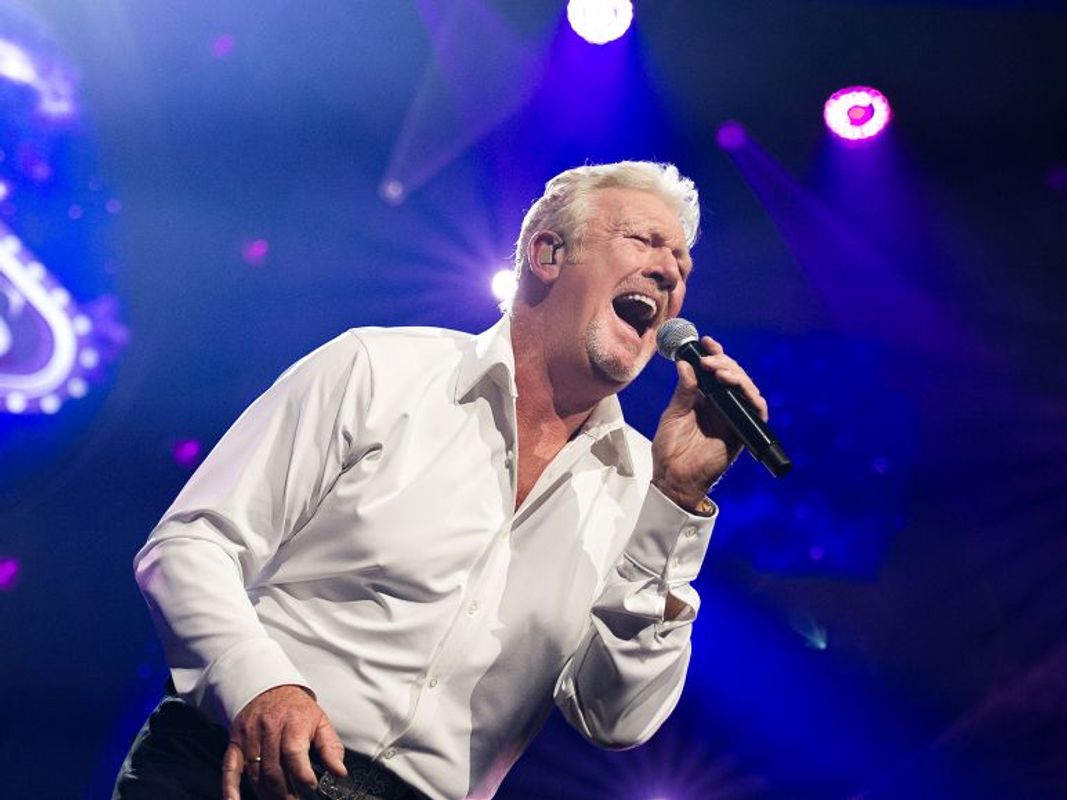 Mr Jones zingt uitbundig door de microfoon die hij vasthoudt. De achtergrond is blauw gekleurd.