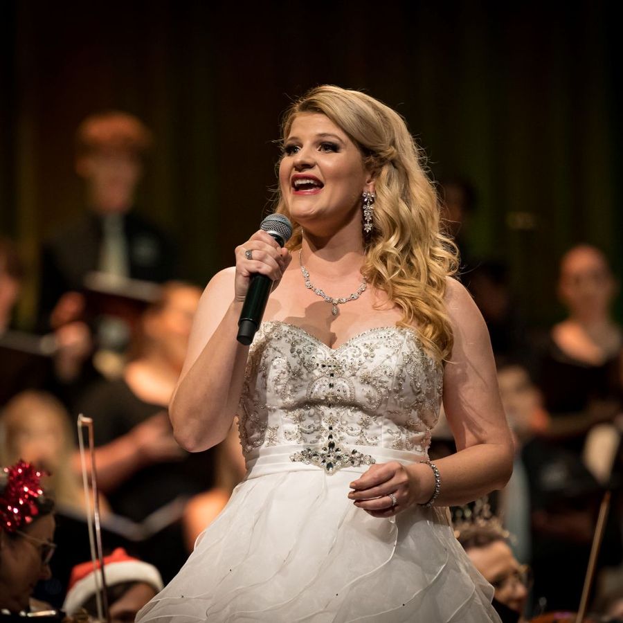 Een vrouw in een witte jurk houdt een microfoon vast en zingt. Op de achtergrond staat een koor.