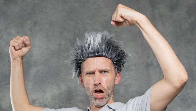Een man met een pruik van grijs haar dat rechtop staat, staat voor een grijze achtergrond met zijn armen omhoog alsof hij spierballen laat zien en kijkt stoer.