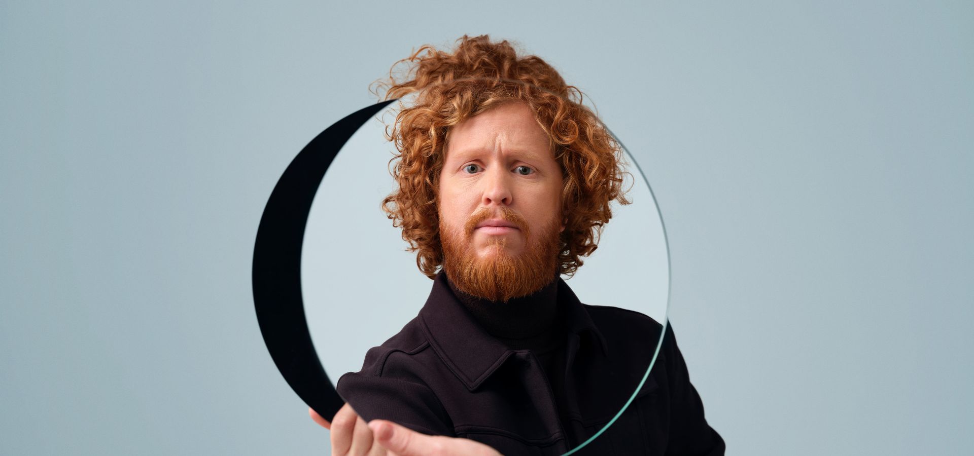 Man in zwart en bos krullen houdt zichzelf een spiegel voor. 