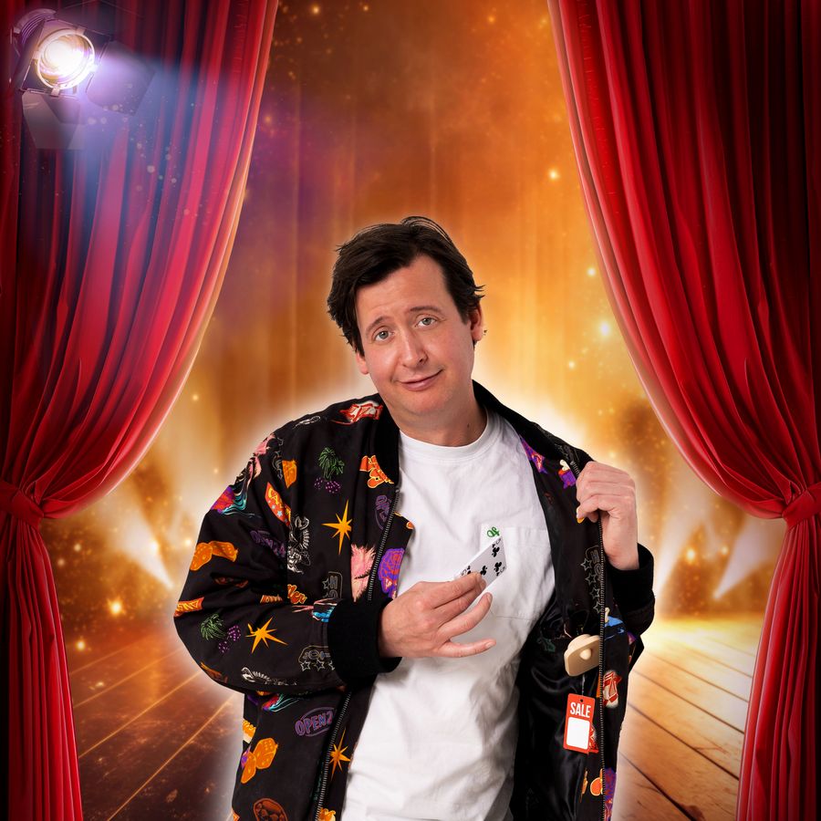 Steven kijkt glimlachend naar de camera en houdt zijn gekleurde jasje half open. Hij staat voor een rood doek die half open zijn en waar een gouden achtergrond te zien is. 